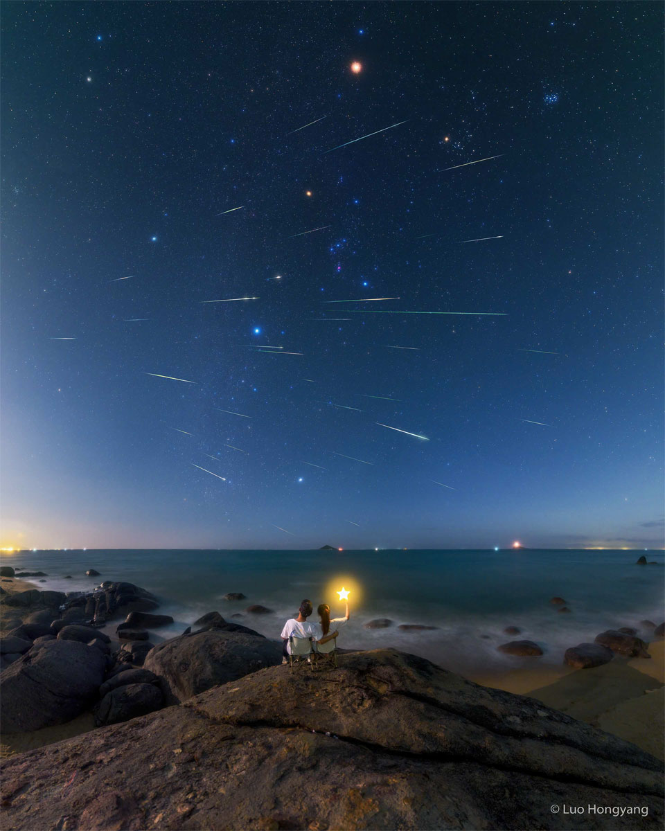 Prezentowane zdjęcie jest złożeniem obrazów wielu śladów meteorów, przecinających niebo w słynnym gwiazdozbiorze Oriona.
Na pierwszym planie widoczna jest dwójka ludzi, siedziących obok siebie i patrzących w niebo. Jedna osoba trzyma różdżkę ze świecącą gwiazdą na jej końcu.
Więcej szczegółowych informacji w opisie poniżej.