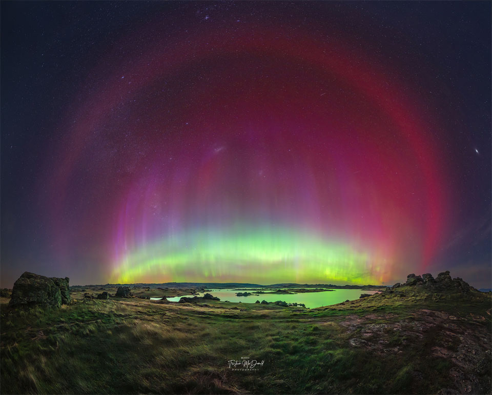 A flat landscape with a pond is imaged at night below
a starfield. A multicolored aurora is seen in an arc across
 the image center. Around this arc is another red arc that
is particularly smooth. 
Więcej szczegółowych informacji w opisie poniżej.